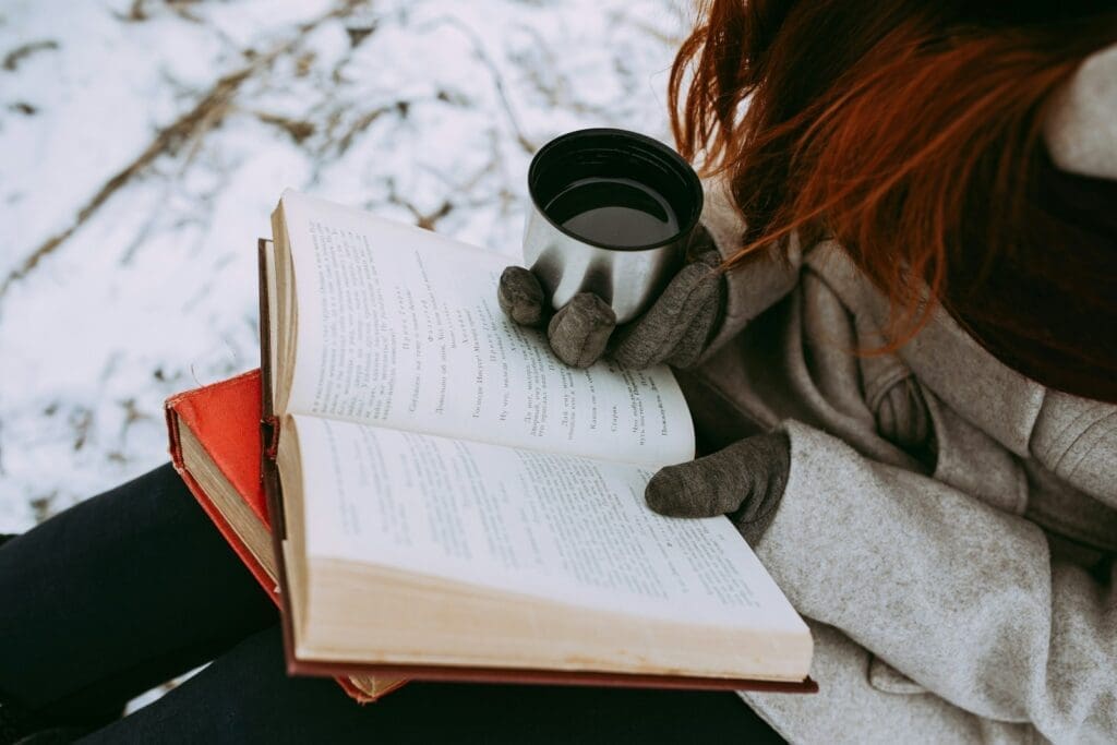 person in gray sweater reading book in the snow, sunlight helps us maintain our winter wellness