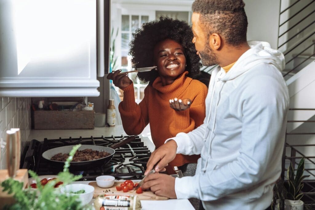 A Black man in white dress shirt cooking with a Black woman in brown long sleeve shirt. Cooking a meal together and reconnecting as a couple can help you beat stress