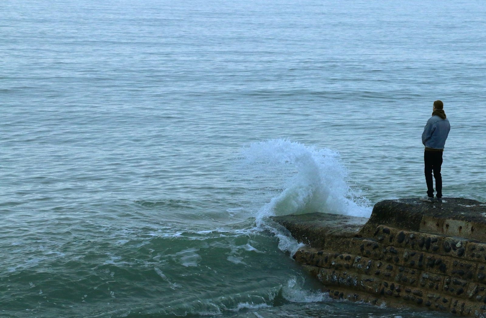brown rock formation on sea during daytime, a lonely figure stands on the rocks looking out