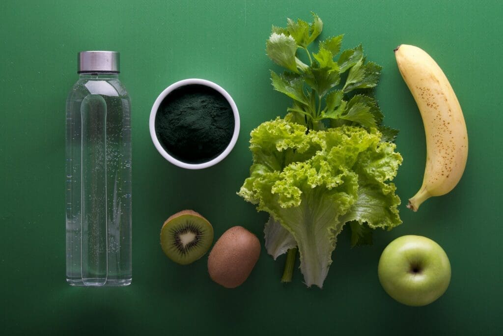 assorted fruits and vegetables on green surface with a bottle if water. These foods have the fiber and hydration key to gut health and digestion.