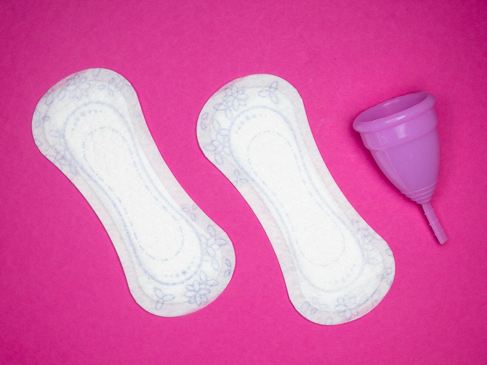 a pair of menstrual pads next to a menstrual cup on a hot pink backgrouund. Naturopathic care for PMS can make your time of the month more bearable