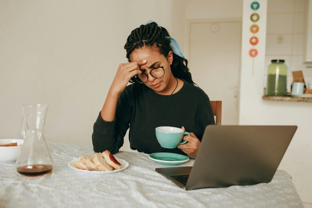 Black woman in black long sleeve shirt sitting and drinking coffee and looking at a laptop