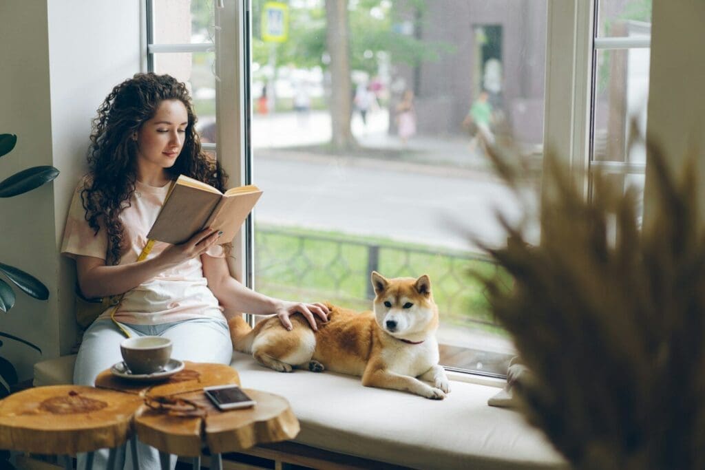 a woman sitting on a window sill reading a book next to a dog. Pets relieve stress a key component to boosting GLP 1 naturally